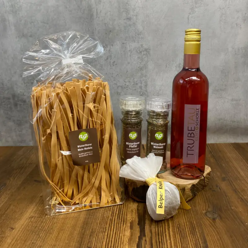 Herbst_Pasta_Gourmetbox Gift set on a wooden table: a large bag of flat pasta, two pepper mills, a round spice bundle tied with a yellow ribbon, and a tall pink bottle of beverage nearby.