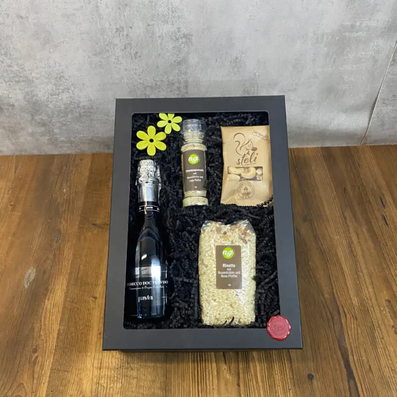 Tete-a-Tete Black windowed gift box showing a bottle of sparkling wine, a spice/pepper grinder, a bag of risotto, crackers, and two yellow flowers on black packing material.